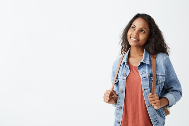 Fröhliche wunderschöne junge afroamerikanische Frau, die Jeansjacke und rotes T-Shirt mit dunklem Haar trägt, träumerisch lächelnd, während sie an etwas Angenehmes denkt. Hübsches Mädchen lässig gekleidet