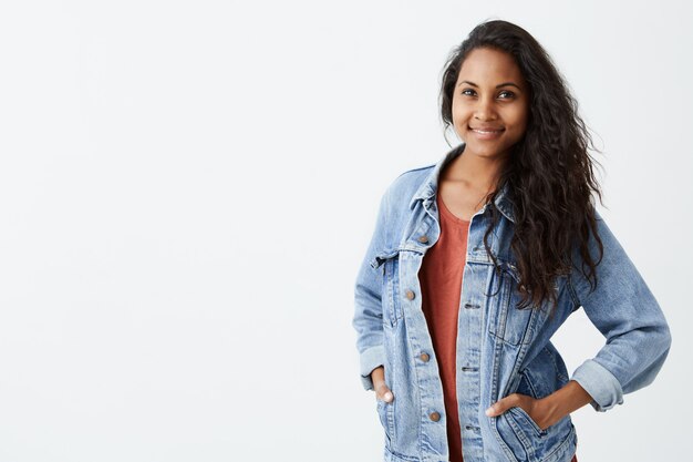 Fröhliche, wunderschöne junge Frau mit dunklem, welligem Haar, die eine Jeansjacke im roten T-Shirt trägt und angenehm lächelt und mit den Händen in den Taschen steht. Hübsches Mädchen, lässig gekleidet.