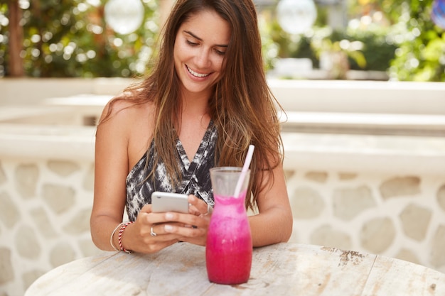 Fröhliche wunderschöne weibliche Dame mit luxuriösem dunklem Haar freut sich über Benachrichtigung auf dem Handy, SMS-Feedback, umgeben von frischem Smoothie, genießt kostenlose Internetverbindung im Café