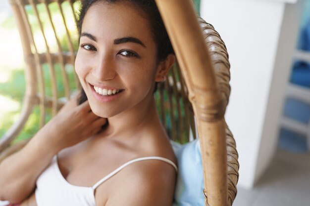 Fröhliche zarte glückliche Frau mit dunklem lockigem Haar, das Ferien genießt, die im Rattanstuhl sitzen