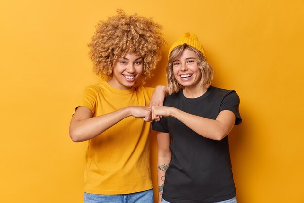 Fröhliche zwei junge Frauen stimmen zu, als Team in lässigem Outfit zu arbeiten, das glücklich isoliert über lebhaftem gelben Hintergrund lächelt. Weibliche Partner genießen Teamarbeit und feiern Erfolge