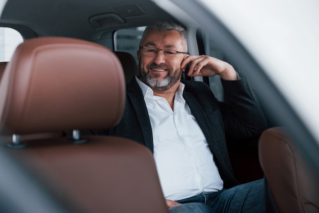 Fröhlicher älterer Geschäftsmann in Brillen, die auf der Rückseite des Autos sitzen und lächeln