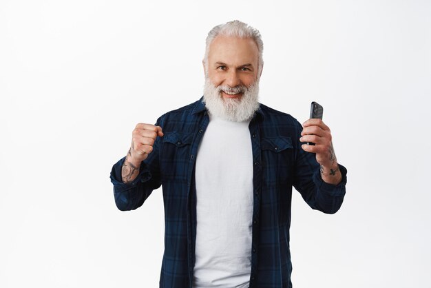 Fröhlicher älterer Mann, der auf dem Handy gewinnt, feiert und lächelt zufrieden, hält Smartphone und ballt triumphierend die Fäuste, erreicht das Ziel, das über weißem Hintergrund steht