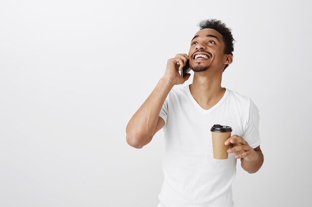Fröhlicher Afroamerikaner, der am Telefon spricht, glücklich lächelt und Kaffee trinkt, aufblickend