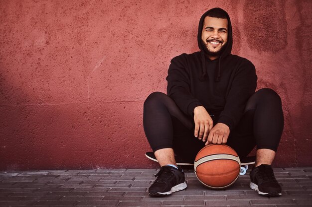 Fröhlicher afroamerikanischer bärtiger Typ in schwarzem Hoodie und Sportshorts, der auf einem Skateboard mit Basketball sitzt und sich an die Wand lehnt.