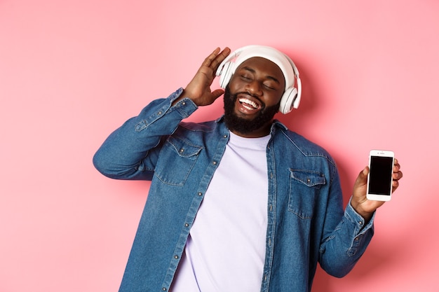 Fröhlicher afroamerikanischer Hipster-Typ, der Musik in Kopfhörern hört, die Telefonbildschirm-App zeigt und mitsingt, auf rosafarbenem Hintergrund steht