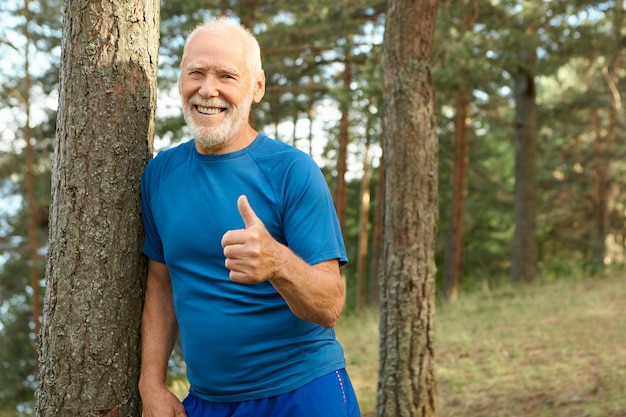 Fröhlicher attraktiver pensionierter Mann mit Glatze und grauem Bart, der draußen in Sportkleidung posiert, die glücklich lächelt, Daumen hoch Geste zeigt, aktiven gesunden Lebensstil wählt, voller Energie