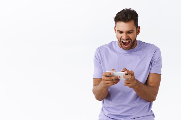 Fröhlicher, aufgeregter, gutaussehender hispanischer, bärtiger Kerl in lila T-Shirt, der erstaunt und begeistert aussieht, wenn er ein tolles neues Smartphone-Spiel spielt, das schreit und lächelt, erfreut, weißen Hintergrund zu gewinnen