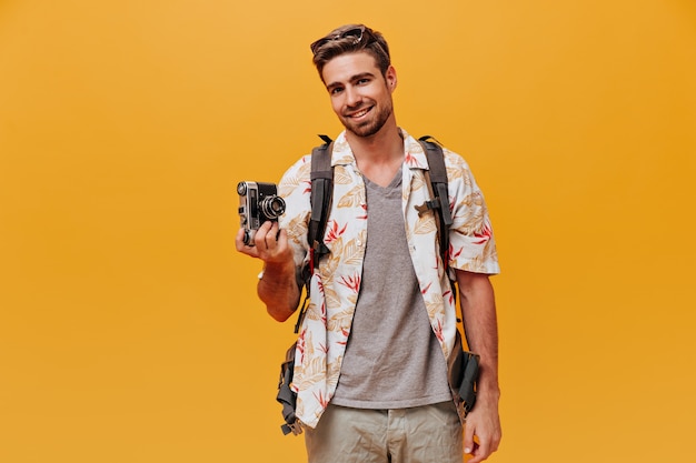 Fröhlicher bärtiger Kerl in grauem T-Shirt und modisch bedrucktem Hemd lächelt und hält Kamera an isolierter orangefarbener Wand