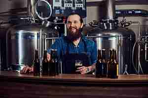 Kostenloses Foto fröhlicher, bärtiger, tätowierter hipster-mann in jeanshemd und schürze, der in einer brauerei arbeitet und hinter einer theke steht, hält ein glas bier zur qualitätskontrolle.