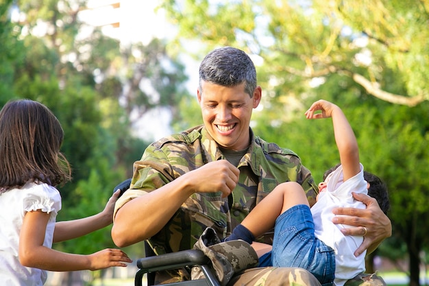 Fröhlicher behinderter Militärvater, der Freizeit mit zwei Kindern im Park genießt. Mädchen, das Rollstuhlgriffe hält, Junge, der auf Papas Schoß ruht. Kriegsveteran oder Behindertenkonzept