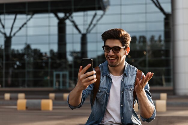 Fröhlicher brünetter Mann mit Sonnenbrille und Jeansjacke lächelt und hält Telefon Schöner Kerl nimmt Selfie nach draußen