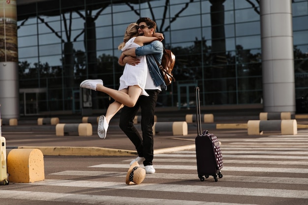 Fröhlicher brünetter Mann umarmt seine Freundin im weißen Kleid Frau trifft Freund am Flughafen Touristische Pose auf Zebrastreifen mit Koffer
