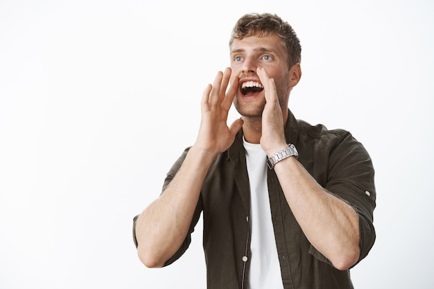 Fröhlicher, charismatischer junger blonder Mann mit Borsten und blauen Augen, der laut schreit, Händchen haltend in der Nähe des geöffneten Mundes, als er in die Ferne ruft und mit Aufregung und Freude lächelt und über grauer Wand posiert