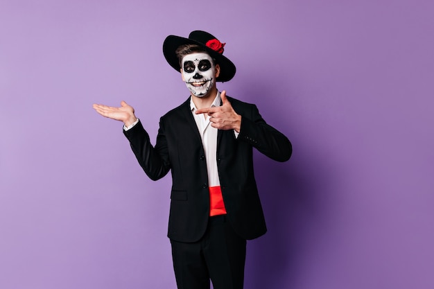 Fröhlicher, enthusiastischer Mann mit gemaltem Gesicht für Halloween zeigt seinen Finger auf Platz für Text auf lila Hintergrund.