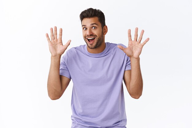 Fröhlicher, enthusiastischer, moderner Typ in lila T-Shirt hat nichts zu verbergen, hebt die Hände zur Hingabe oder Wiederbehandlung, lächelt freudig, winkt mit den Händen in hallo, freundliche Grußgeste, weiße Wand