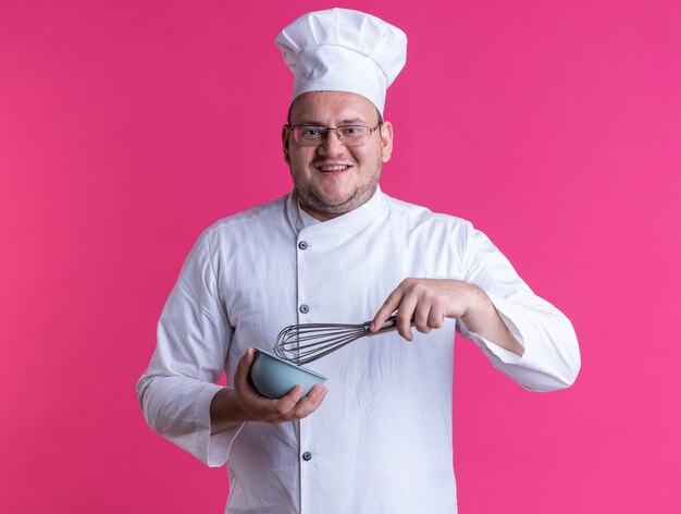 Fröhlicher erwachsener männlicher Koch mit Kochuniform und Brille mit Schneebesen und Schüssel mit Blick auf die Kamera isoliert auf rosa Hintergrund