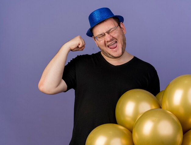 Fröhlicher erwachsener slawischer Mann in optischer Brille mit blauem Partyhut hebt die Faust im Stehen mit Heliumballons