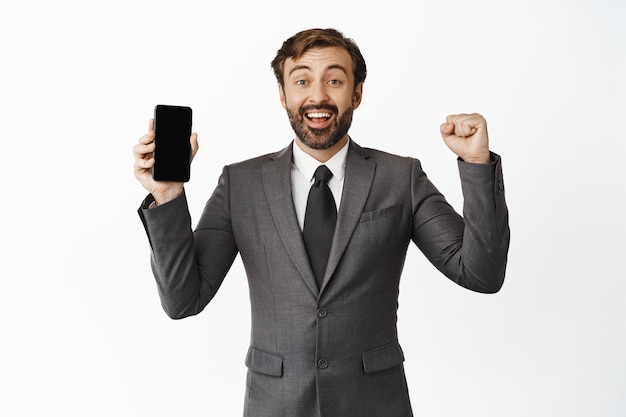 Fröhlicher Firmenmann, der sich freut, den Bildschirm des Mobiltelefons zu zeigen und vor Freude zu springen, zufrieden prahlt und mit dem weißen Hintergrund des Gewinns prahlt