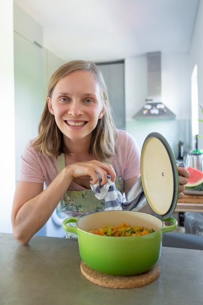 Fröhlicher Food-Blogger, der Topf mit Gemüsemehl öffnet, sich auf Tisch stützt, für Kamera posiert und lächelt. Vertikaler Schuss. Kochen zu Hause Konzept