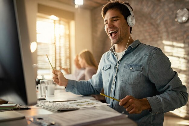 Fröhlicher Freiberufler, der Spaß hat, während er am Schreibtisch arbeitet und Musik über Kopfhörer hört. Seine Kollegen sind im Hintergrund