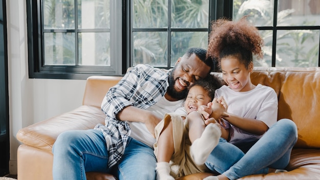 Fröhlicher, fröhlicher afroamerikanischer Familienvater und Tochter, die beim Geburtstag im Haus Spaß beim Kuscheln auf dem Sofa haben.