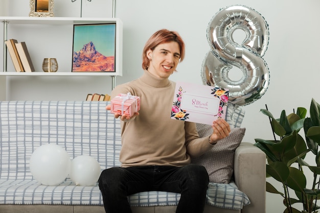 Fröhlicher, gutaussehender Kerl am glücklichen Frauentag, der ein Geschenk mit einer Grußkarte auf dem Sofa im Wohnzimmer hält