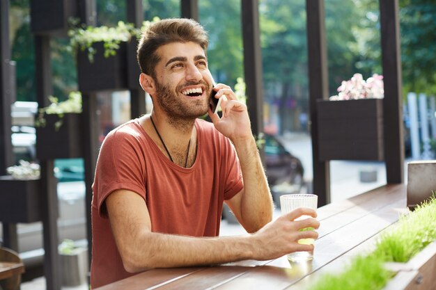 Fröhlicher gutaussehender Mann, der lacht und lächelt, während er auf Handy vom Außencafé spricht