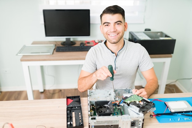 Fröhlicher gutaussehender Mann, der lächelt und Augenkontakt herstellt, während er einen Schraubendreher hält, um einen kaputten CPU-Computer in seinem Geschäft zu reparieren