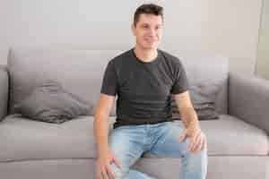 Kostenloses Foto fröhlicher gutaussehender mann, der lässiges t-shirt und jeans trägt, zu hause auf der couch sitzt, hände auf dem schoß hält, wegschaut und lächelt. männliches porträtkonzept