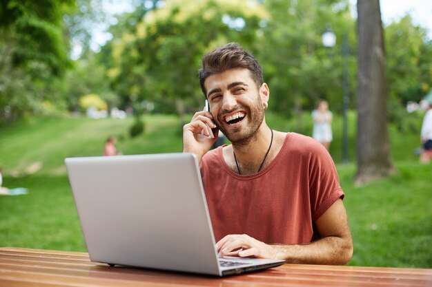 Fröhlicher gutaussehender Mann, der Laptop im Park benutzt und glücklich am Telefon spricht