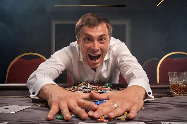 Kostenloses Foto fröhlicher gutaussehender mann in einem weißen hemd spielt poker am tisch im casino. er freut sich über den gewinn, nimmt die chips und blickt mit offenem mund in die kamera. glücksspiel um geld. spiele von