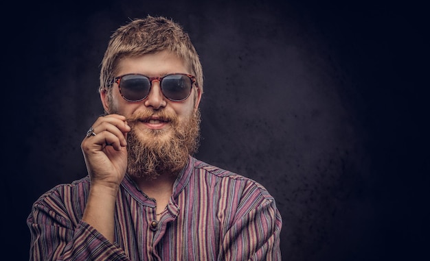 Fröhlicher Hipster-Typ mit Sonnenbrille in einem altmodischen Hemd korrigiert seinen Schnurrbart. Getrennt auf einem dunklen Hintergrund.