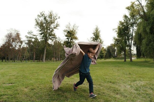 Fröhlicher Junge, der mit einer fliegenden Decke im Park läuft