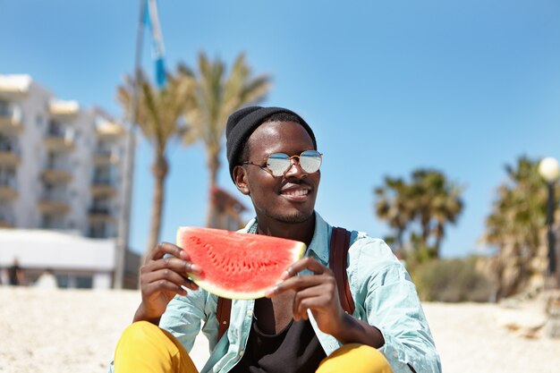 Fröhlicher junger Afroamerikaner, gekleidet in trendige Kleidung, die schöne Zeit draußen am Meer hat, reife saftige Wassermelone und gutes sonniges Wetter genießt, breit lächelt und schöne Seelandschaft bewundert