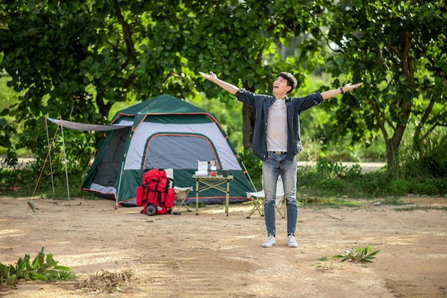 Fröhlicher junger Backpacker-Mann, der vor dem Zelt im Wald mit Kaffeeset steht und offene Arme hat und während des Campingausflugs im Sommerurlaub frische Kaffeemühle zubereitet