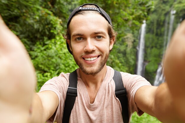 Fröhlicher junger bärtiger Hipster, der Baseballmütze trägt, die Selfie in seinen Ferien im exotischen Land nimmt