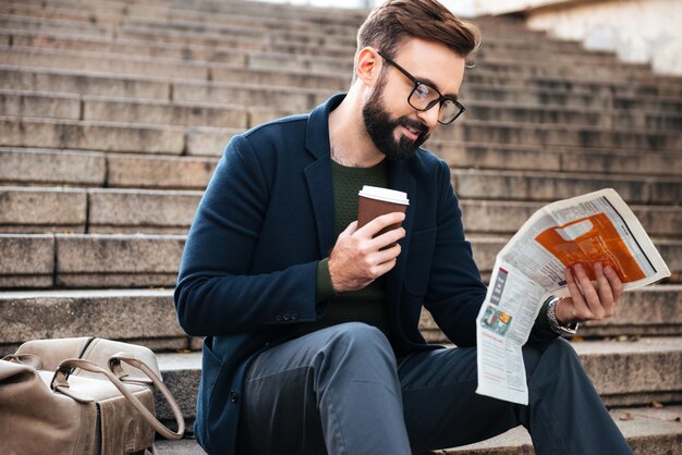 Fröhlicher junger bärtiger Mann, der draußen auf Stufen sitzt, die Zeitung lesen