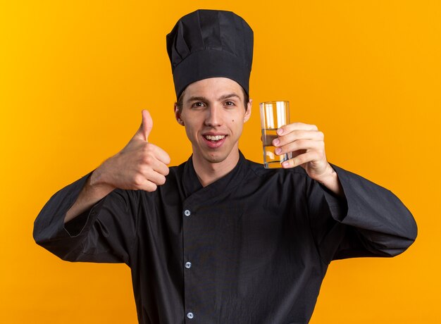 Fröhlicher junger blonder männlicher Koch in Kochuniform und Mütze mit Glas Wasser mit Daumen nach oben