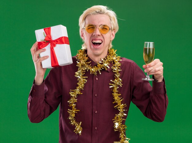 Fröhlicher junger blonder Mann mit Brille mit Lametta-Girlande um den Hals, der ein Glas Champagner und ein Geschenkpaket hält und auf die Kamera schaut, die isoliert auf grünem Hintergrund schreit