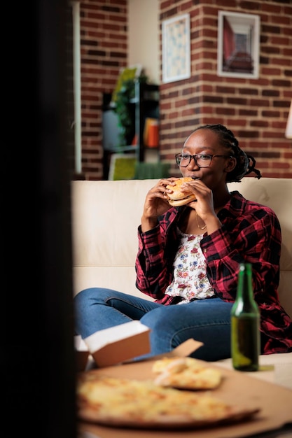 Fröhlicher junger Erwachsener, der Burger isst und eine Flasche Bier trinkt, Freizeitaktivitäten und lustige Filme im Fernsehen genießt. Fast-Food-Lieferservice zum Mitnehmen mit Getränken und Snacks zu Hause.