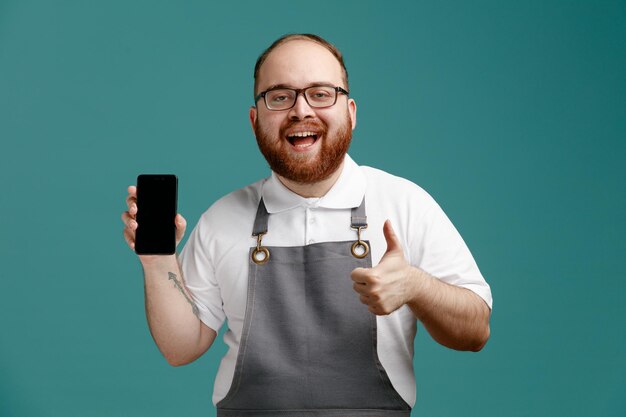 Fröhlicher junger friseur in uniform und brille, der das handy zeigt, das in die kamera schaut und den daumen isoliert auf blauem hintergrund zeigt