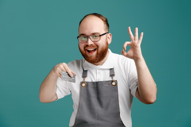 Fröhlicher junger Friseur in Uniform und Brille mit Kreditkarte, der in die Kamera schaut und ein Ok-Zeichen auf blauem Hintergrund zeigt