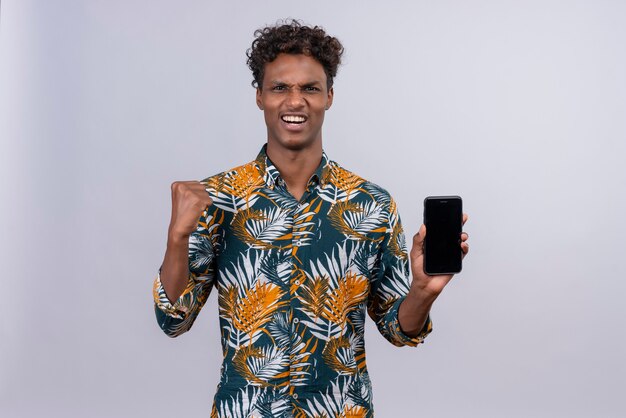 Fröhlicher junger gutaussehender dunkelhäutiger Mann mit lockigem Haar im Blatt bedruckten Hemd, das Smartphone mit geballter Faust hält