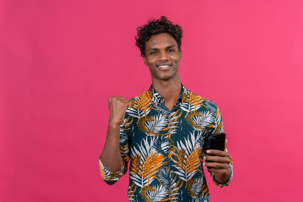 Fröhlicher junger gutaussehender dunkelhäutiger Mann mit lockigem Haar im Blatt bedruckten Hemd, das Smartphone mit geballter Faust hält