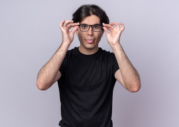 Fröhlicher junger gutaussehender Kerl mit schwarzem T-Shirt, das eine Brille trägt und hält, die Zunge isoliert auf weißer Wand zeigt