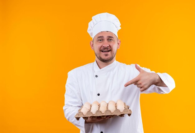 Fröhlicher junger gutaussehender Koch in Kochuniform, der auf einen Karton mit Eiern zeigt, die auf orangefarbener Wand isoliert sind?