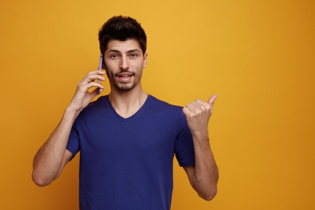 Fröhlicher junger gutaussehender Mann, der am Telefon spricht und in die Kamera blickt, die auf gelbem Hintergrund zur Seite zeigt