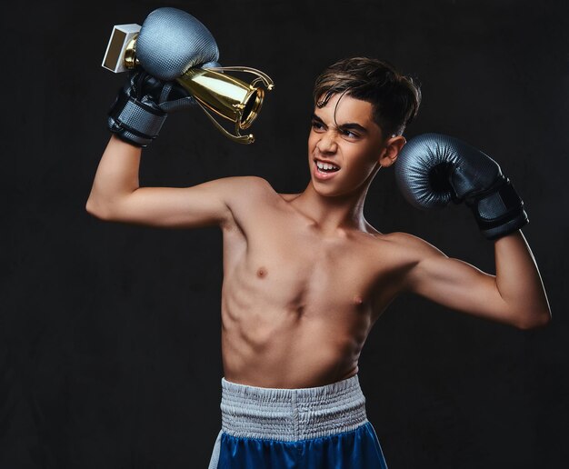 Fröhlicher junger, hemdloser Boxer-Champion mit Handschuhen hält einen Siegerpokal. Getrennt auf einem dunklen Hintergrund.