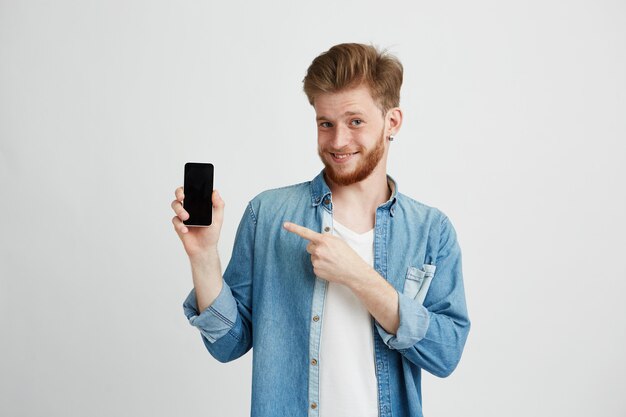Fröhlicher junger hübscher Mann, der lächelnden Zeigefinger auf Smartphone in seiner Hand über weißem Hintergrund zeigt.